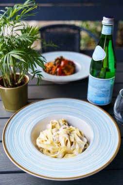 Mavi çizgili seramik tabakta servis edilen zengin ve kremsi peynir soslu fettuccine alfredo makarnası. Tabak koyu ahşap bir masada, başka bir kaplama ve yeşil bitkilerle birlikte..