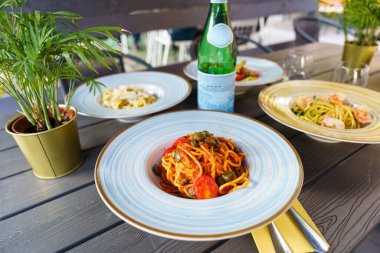 Renkli İtalyan makarna tabakları ve bir şişe köpüklü su ile açık hava masası taze ve davetkar bir Akdeniz yemek atmosferi yaratıyor..