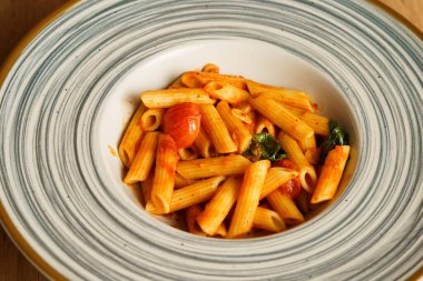 Canlı domates sosuyla kaplanmış, taze kiraz domatesleri ve fesleğen yapraklarıyla süslenmiş minimalist bir makarna servisi. Sıcak ahşap arka plan bu İtalyan yemeğinin tazeliğini vurguluyor..