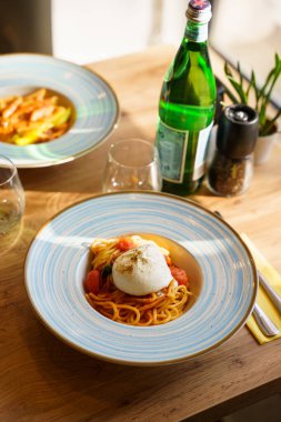 Üzerine burrata peyniri konmuş spagetti makarnası. Kiraz domatesli ve taze fesleğenli domates sosuyla servis edilmiş. Doğal ışık altında bir İtalyan yemeği..