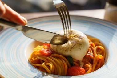 Domates soslu ve kiraz domatesli spagettiye yerleştirilmiş kremalı burata peynirini dilimleyen çatal ve bıçağın yakın plan görüntüsü zengin dokuları ve çekici aromayı yakalıyor..