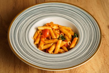 Canlı domates sosuyla kaplanmış, taze kiraz domatesleri ve fesleğen yapraklarıyla süslenmiş minimalist bir makarna servisi. Sıcak ahşap arka plan bu İtalyan yemeğinin tazeliğini vurguluyor..