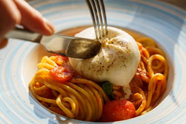 Domates soslu ve kiraz domatesli spagettiye yerleştirilmiş kremalı burata peynirini dilimleyen çatal ve bıçağın yakın plan görüntüsü zengin dokuları ve çekici aromayı yakalıyor..