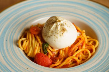 Bir burrata topuyla kaplı bir spagetti tabağı, kiraz domatesli ve fesleğenli domates sosuyla servis ediliyor. Kremalı peynir, canlı makarnayla çok güzel zıtlaşıyor..