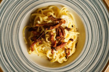 Çizgili seramik tabakta gevrek pancetta ile kaplanmış güzel kaplanmış bir spagetti carbonara. Kremalı sos ve altın renkler sıcak ve kırsal bir İtalyan yemeği deneyimi yaratır..