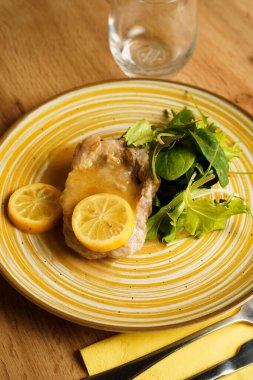 Limon dilimleri ve hafif sosla servis edilen yumuşak domuz pirzolası ve yanında taze yeşil salata. Tabak, ahşap bir masanın üzerinde kırsal sarı çizgili seramik tabakta sunuluyor..