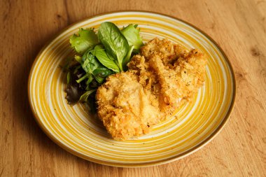 Sarı çizgili tabakta canlı karışık salata sebzeleriyle servis edilen kızarmış tavuk filetoları. Tabak, taze, yapraklı elementlerle çıtır çıtır dokuya zıt düşüyor..