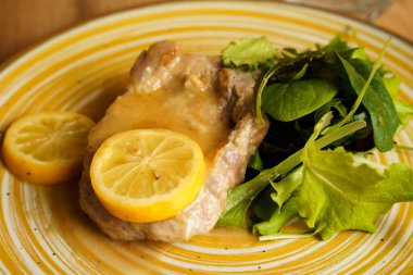 Limon dilimleri ve hafif sosla servis edilen yumuşak domuz pirzolası ve yanında taze yeşil salata. Tabak, ahşap bir masanın üzerinde kırsal sarı çizgili seramik tabakta sunuluyor..