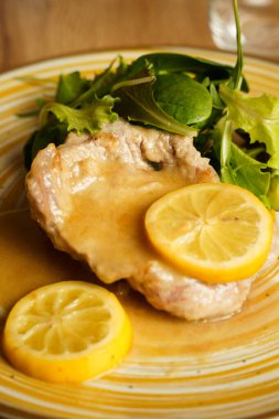 Limon dilimleri ve hafif sosla servis edilen yumuşak domuz pirzolası ve yanında taze yeşil salata. Tabak, ahşap bir masanın üzerinde kırsal sarı çizgili seramik tabakta sunuluyor..