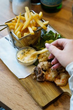 Bir el patates kızartmasını tartar sosuna batırır yanında çıtır deniz ürünleri, sepette patates kızartması ve taze yeşillikler, gazete tarzı bir kalasla ahşap bir tahtaya dizilir..