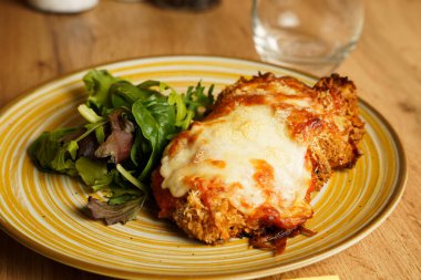 Eritilmiş peynir ve domates soslu fırında tavuk göğsü. Kırsal sarı çizgili tabakta taze yeşil salata ile servis ediliyor..