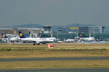 Fotoğrafta Frankfurt Havaalanı 'nda önlük sergilenirken, ön planda birkaç yolcu uçağı yer alıyor. Arka planda, terminal binası ve ikonik kontrol kulesi görünür.