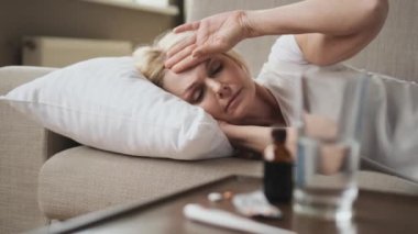 Hasta kadın, baş ağrısı şikayetiyle yatakta yatıyor. Ön taraftaki bir masada, tıbbi tedavisinden kalma haplar var.