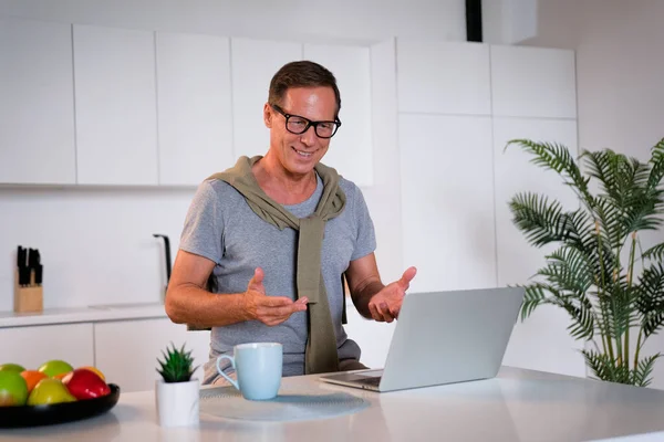 mature man using laptop, making video call, sitting at table in kitchen, senior teacher mentor wearing glasses engaged online conference, recording webinar, teaching