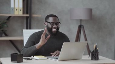 Merhabalar. Genç, Afro-Amerikalı adam kameraya el sallarken iş yerindeki iş yerindeki ofisinde günlük kıyafetleriyle görüntülü arama yapıyor.