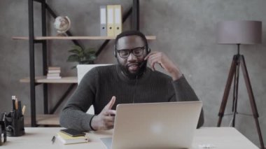 Kulaklık takmış Afrikalı Amerikalı bir erkek dizüstü bilgisayarla online danışmanlık yapıyor. Kulaklıklı adam internette web sitesi yönetiyor..