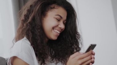 Mutlu milenyal Afrikalı Amerikalı kadın telefon e-ticaret uygulamaları kullanarak akıllı telefon tutarak, mobil oyunlar oynarken, haberci veya sosyal medya ağları sohbet, internette sörf veya manifatura mesaj telefonda