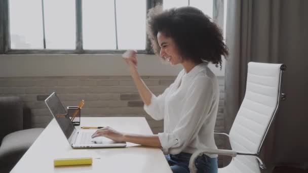 喜びに満ちたアフリカ系アメリカ人の若い女性は ラップトップで自宅の机を見て座って 興奮した出産の男性は コンピュータ上で良いニュースを読んで陶酔感の勝利を感じる 報酬の概念 — ストック動画