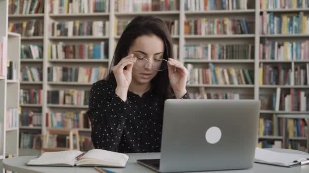 Zmęczona Kobieta Ręką Masuje Oczy Siedząc Przy Stole Tle Biblioteczki — Wideo stockowe