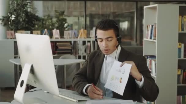 ビジネスマンは自分のコンピュータ上の文書を介して作業し オンラインビデオ会議 ヘッドセットを持つラテン系の男で書類の統計を示しています — ストック動画