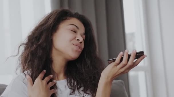 集中笑顔アフリカ系アメリカ人の若い女性は スピーカーで携帯電話で話して自宅でソファに座って 携帯電話で音声メッセージを記録 — ストック動画