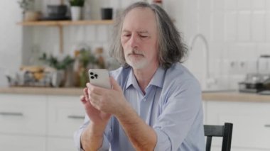 Mutfakta elinde akıllı bir telefonla oturan yaşlı bir adam mesaj atıyordu. ciddi yetişkin emekliler telefonla anında haberci kullanırlar.