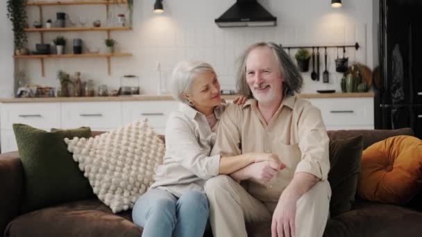 笑顔の高齢者の肖像60代の夫と妻座っていますリラックスします ソファ抱擁抱っこ 幸せな成熟したシニアカップル休息上のソファの上にリビングルームで抱擁見てカメラショー愛とケア — ストック動画