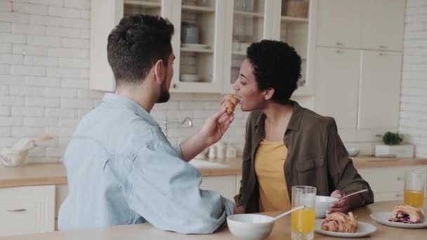 キッチンに座っている間に 幸せな多民族の若いカップルがお互いにクロワッサンを食べます — ストック動画