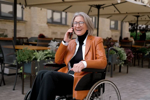 Tekerlekli sandalyedeki yaşlı, kır saçlı kadın dışarıda telefonla konuşuyor..