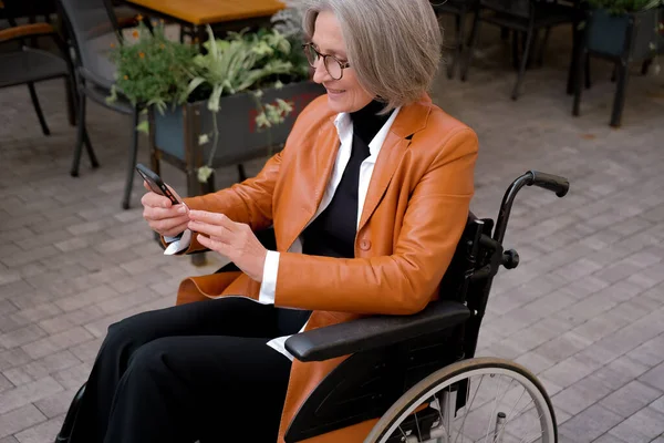 elder woman in wheelchair holding mobile phoneoutdoor. elderly female texting message, using app with cellphone in park.