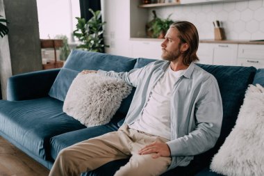 Sarışın, yakışıklı, sakallı bir adam yan bakıyor, rahat giysiler içinde bir kanepeye kıvrılıyor..