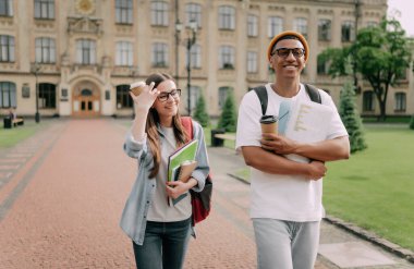 İki uluslararası stil öğrencisi kampüsün etrafında yürüyüp gülümsüyor. Dost canlısı Afrikalı Amerikalı adam ve Kafkas kız üniversitenin yakınında birlikte yürüyorlar.