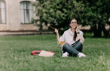 Genç kız öğrenci kampüste çimlerde oturuyor ve neşeli bir şekilde akıllı bir telefonla konuşuyor ve gülümsüyor..
