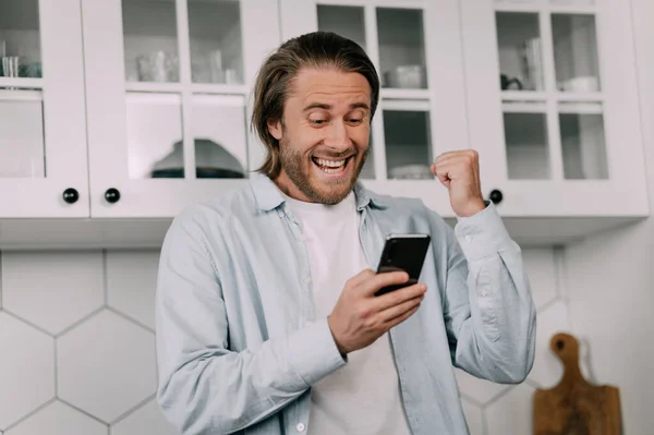 若い男が自宅でスマートフォンを見て成功を喜びます ラッキーベット 株式市場 暗号市場 — ストック写真