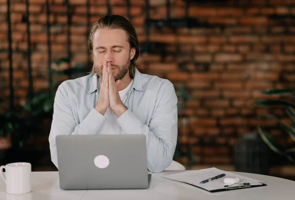 Başarı için dua ediyorum. Düşünceli genç adam çenesini tutmuş ve ofisteki iş yerinde otururken laptopa bakıyor..