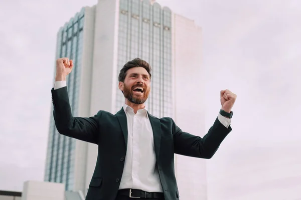 Mutlu, mutlu, heyecanlı Avrupalı, takım elbiseli sakallı iş adamı başarılı bir anlaşma yapıyor ya da kazanıyor, modern ofis binasının yakınında el kaldırıyor, açık havada. Menajer, zafer duyguları, jestler.