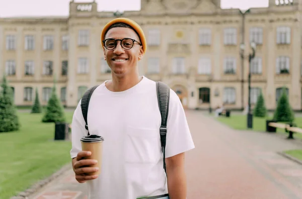 アフリカ系アメリカ人のスタイリッシュなトレンディの学生の肖像で 背景には大学と笑顔でコーヒーを片手にしています — ストック写真