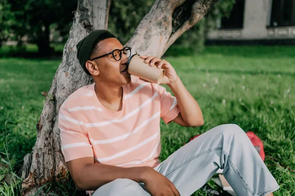 アフリカ系アメリカ人の若者が芝生の上の公園に座ってクラフトカップからコーヒーを飲んで — ストック写真
