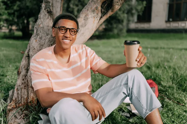 アフリカ系アメリカ人の若者が芝生の上の公園に座ってクラフトカップからコーヒーを飲んで — ストック写真