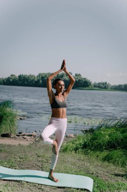 Nehir kıyısında yoga dengesi egzersizi yapan sağlıklı genç bir kadının tam boy portresi..
