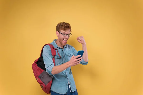 感情的な魅力的な学生は彼の手にスマートフォンを保持し 喜びます 彼の手にスマートフォンを持つ若い男は 黄色の背景に隔離された成功を喜びます コピースペース — ストック写真