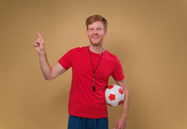 male coach with a soccer ball and a whistle points a finger to an empty space on a beige background