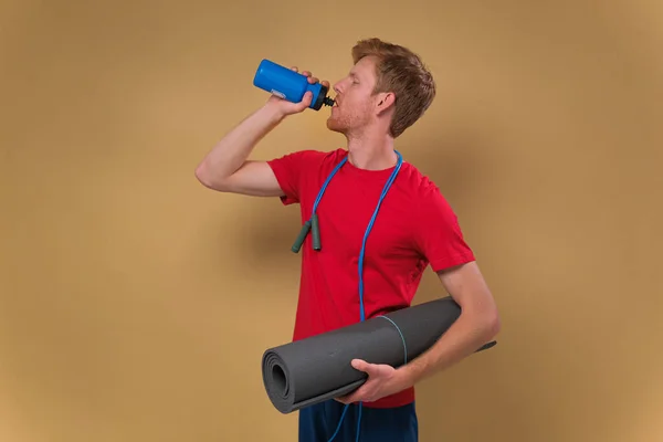 Spor kıyafetleri içinde yoga minderiyle yakışıklı bir adam bej arka planda içme suyu içiyor.