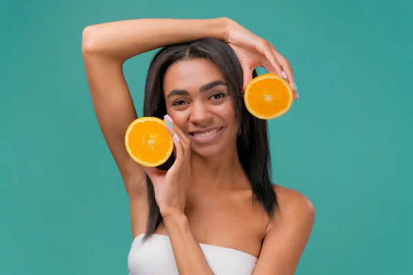 Delighted African American female with showing halves of fresh juicy orange while representing positive aspects of vitamin C for health and beauty on turquoise background