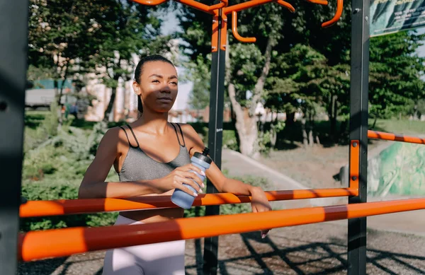 Atletik Afrikalı Amerikalı kadın elinde bir şişe suyla oyun parkında. Su dengesi kavramı.
