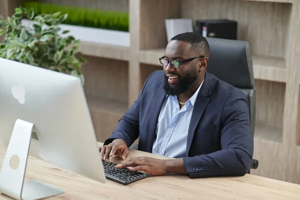 Ofiste çalışırken bilgisayarda daktilo kullanan konsantre Afro-Amerikan erkek işadamı. Yetişkin şirket patronu masasında oturuyor. Modern başarılı adam kavramı. İş liderliği ve yönetim fikri