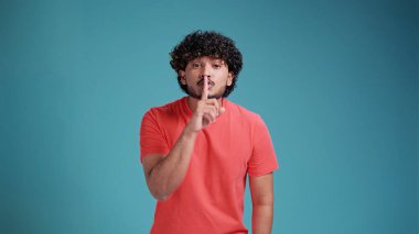 Sessiz olun lütfen. Genç Hintli adam işaret parmağını dudaklarına bastırıyor sessizlik işareti yapıyor. Gizli saklı konuşmayın, dedikoduyu kesin. Mercan tişörtü giyen yakışıklı, sakallı Hintli adam mavi bir arka planda izole edilmiş.