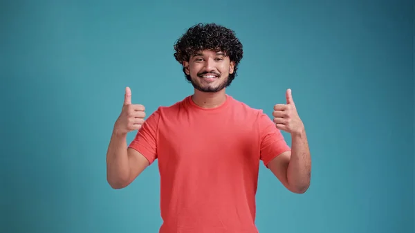 肯定的なインドの若い男がウインクし 親指を表示します 親指を立てて 青いスタジオの背景に隔離された基本的なサンゴのTシャツでカメラを見ているハンサムな成功した男 成功の概念 — ストック写真