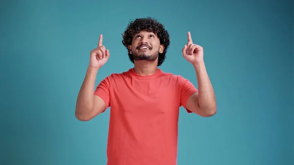 Mercan tişörtü giyen genç Hintli adam mavi stüdyo arka planında izole edilmiş. Neşeli ve yüzünde mutlu ve doğal bir ifadeyle elleri ve parmaklarıyla işaret eden bir gülümsemeyle.