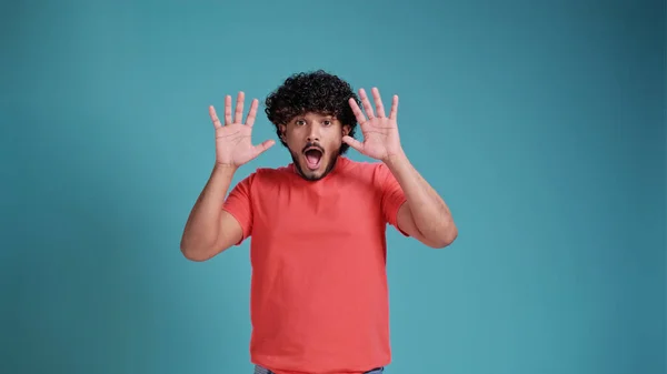 ホラー スリラー 怖い話 青いスタジオの背景にサンゴのTシャツに恐怖の顔を持つ若いおびえたインド人男性 男叫びと手を上げる肖像画 人間の感情 心理状態の概念 — ストック写真